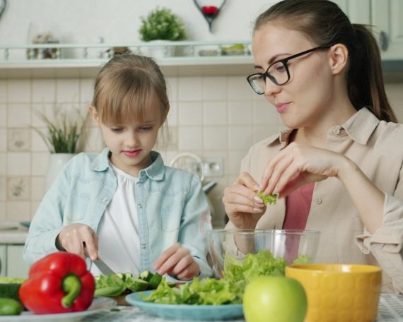 Cute loving daughter is helping mother with salad cutting cucumbers feeding young woman and laughing having fun at home. People and cookery concept.